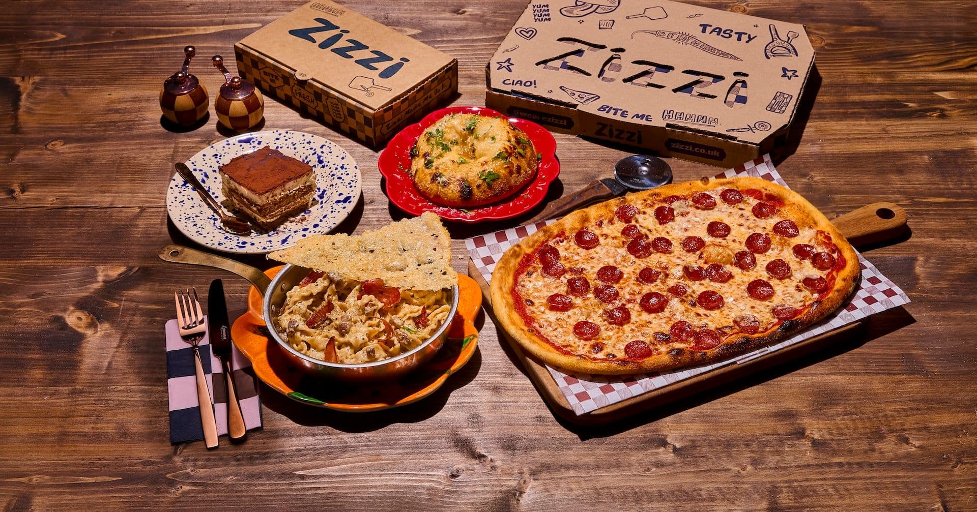 Feast shot of pizza, pasta, tiramisu and garlic bombe, with takeaway boxes in background