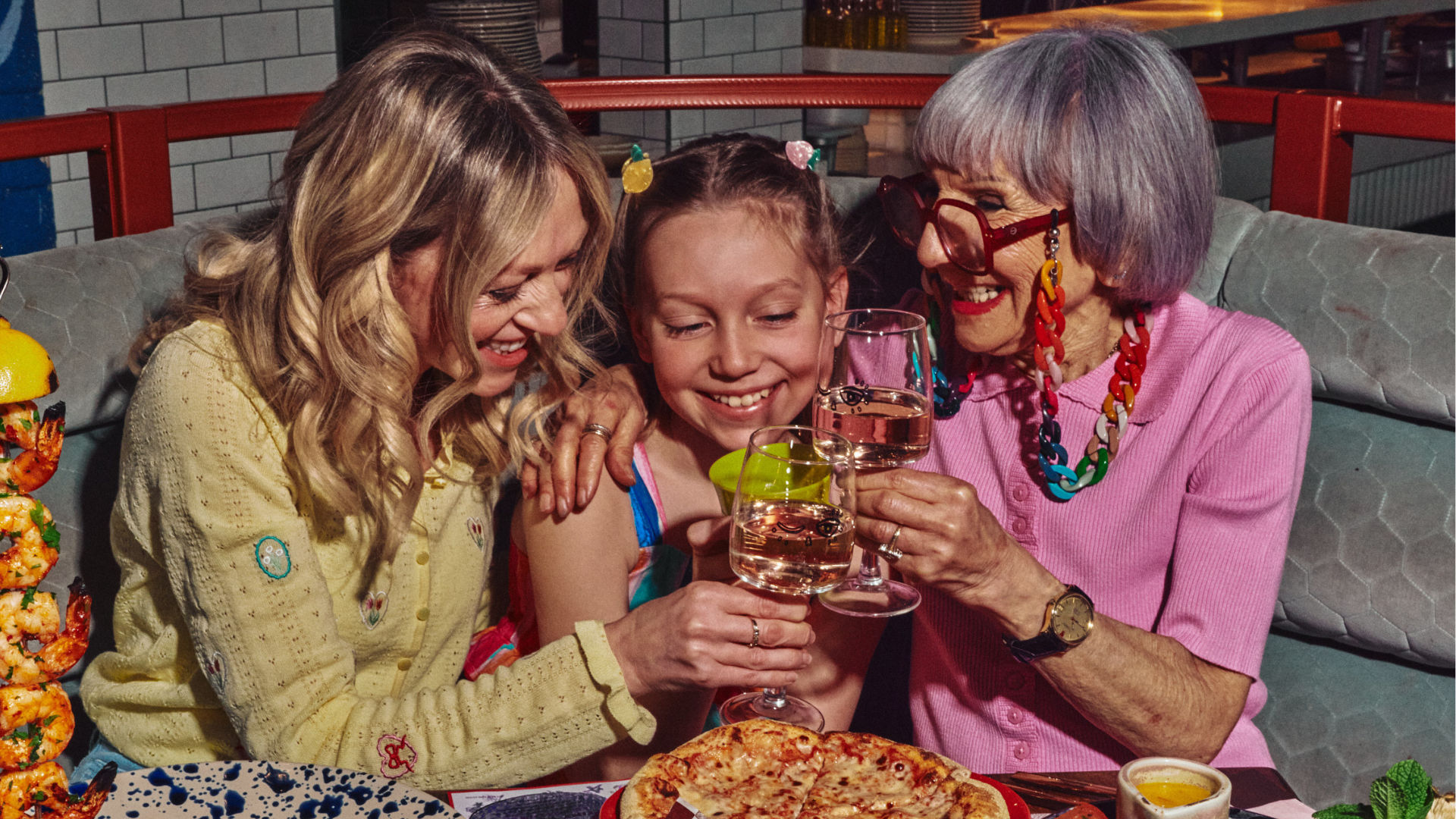 Grandmother, mother and daugher smiling at the table with a table of food and drink. 